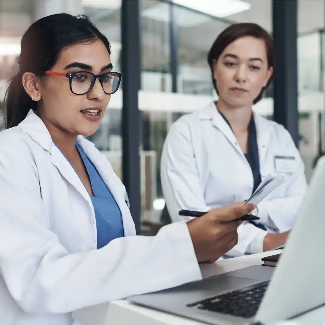 Two female physicians in white coats collaborating at a laptop in a clinical office setting.