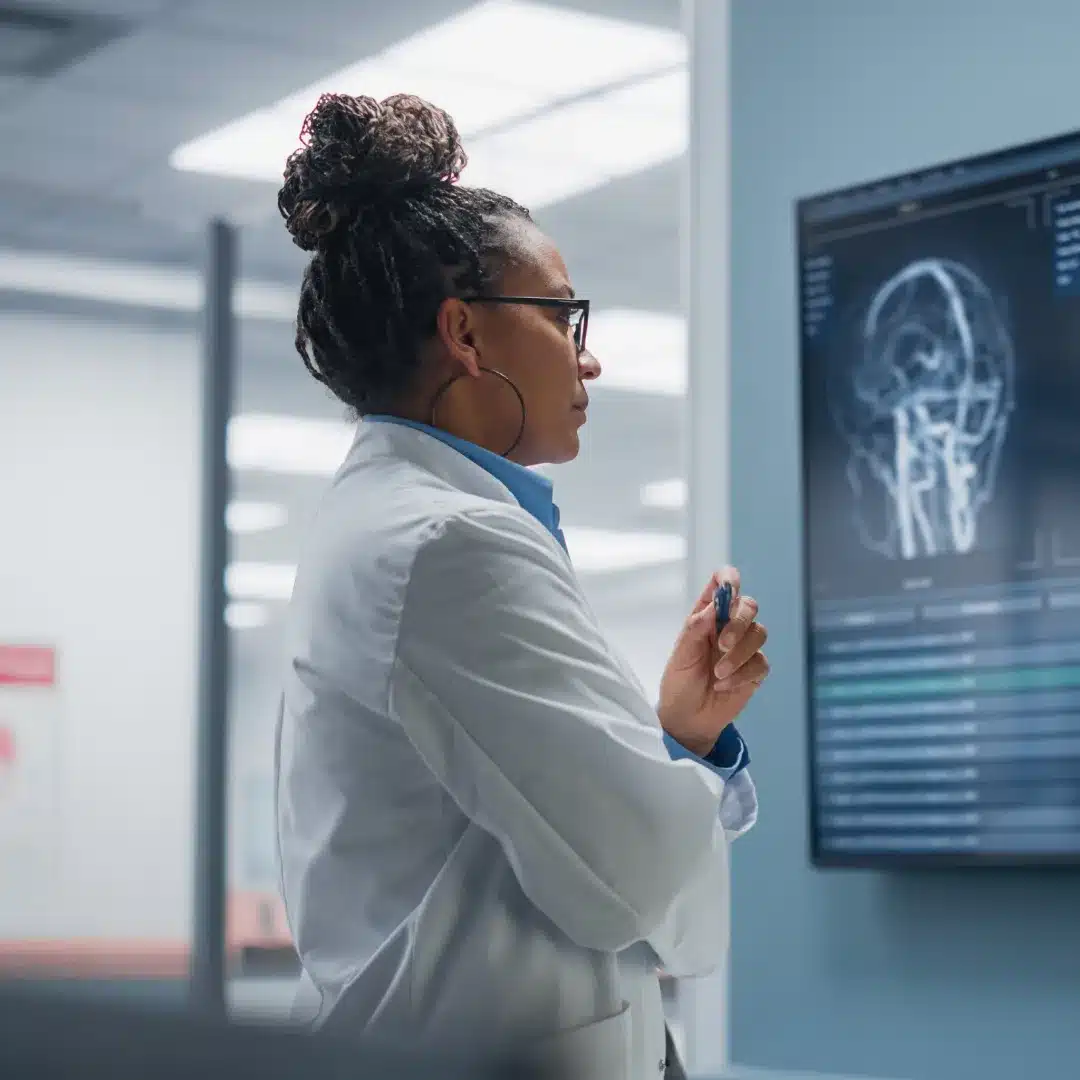 Female physician in a white coat studying a brain scan on a wall-mounted monitor in a clinical lab.