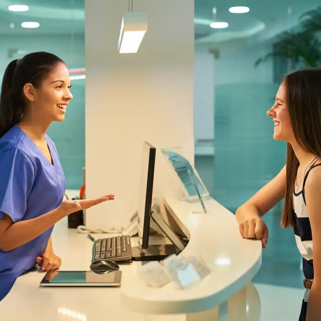 Nurse in dark blue scrubs greeting a patient at a modern medical reception desk with a tablet nearby.