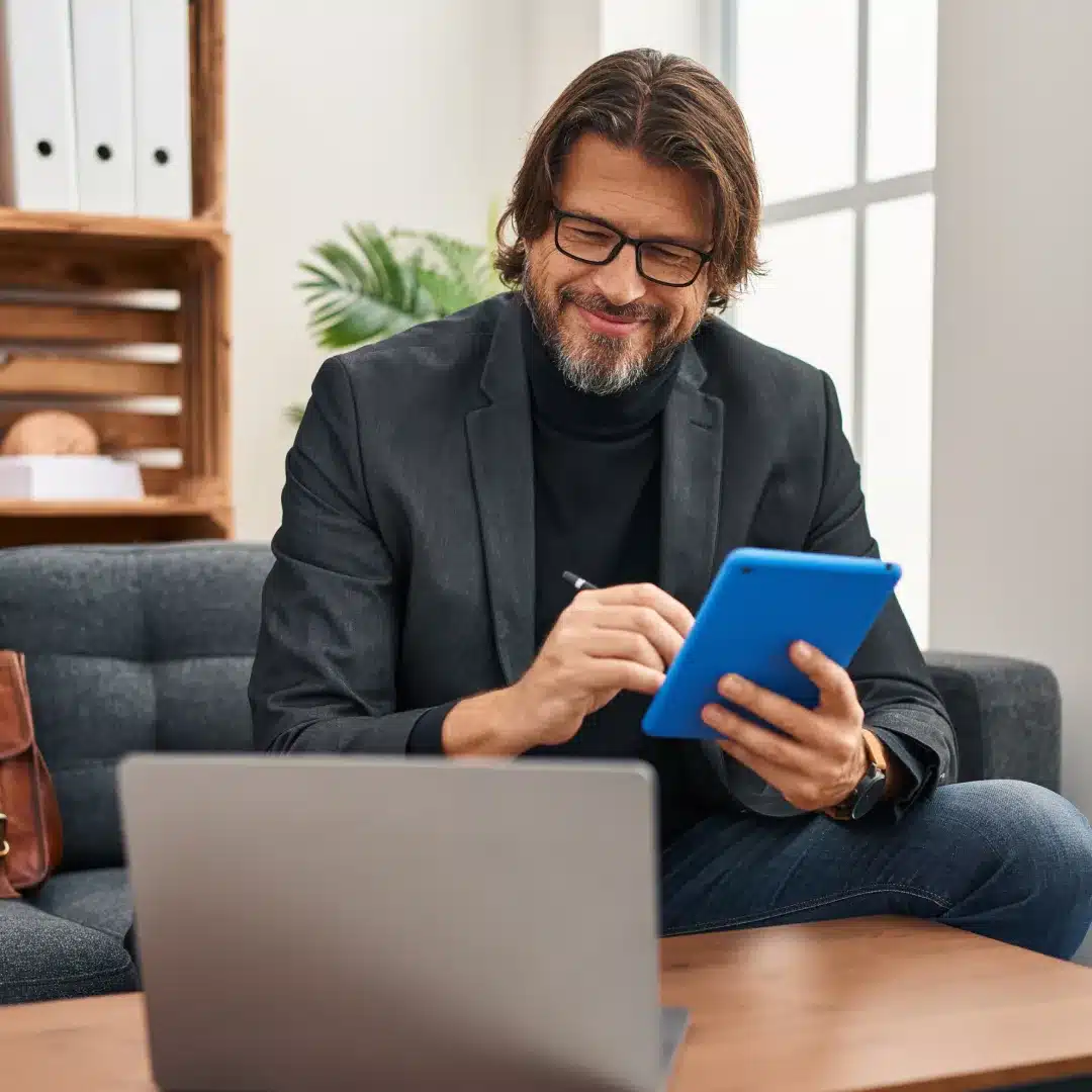 Man in a dark blazer and glasses writing on a tablet with a stylus while seated beside a laptop.