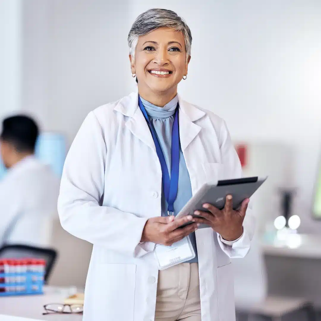 Senior female physician in a white coat and badge holding a tablet in a bright clinical office.