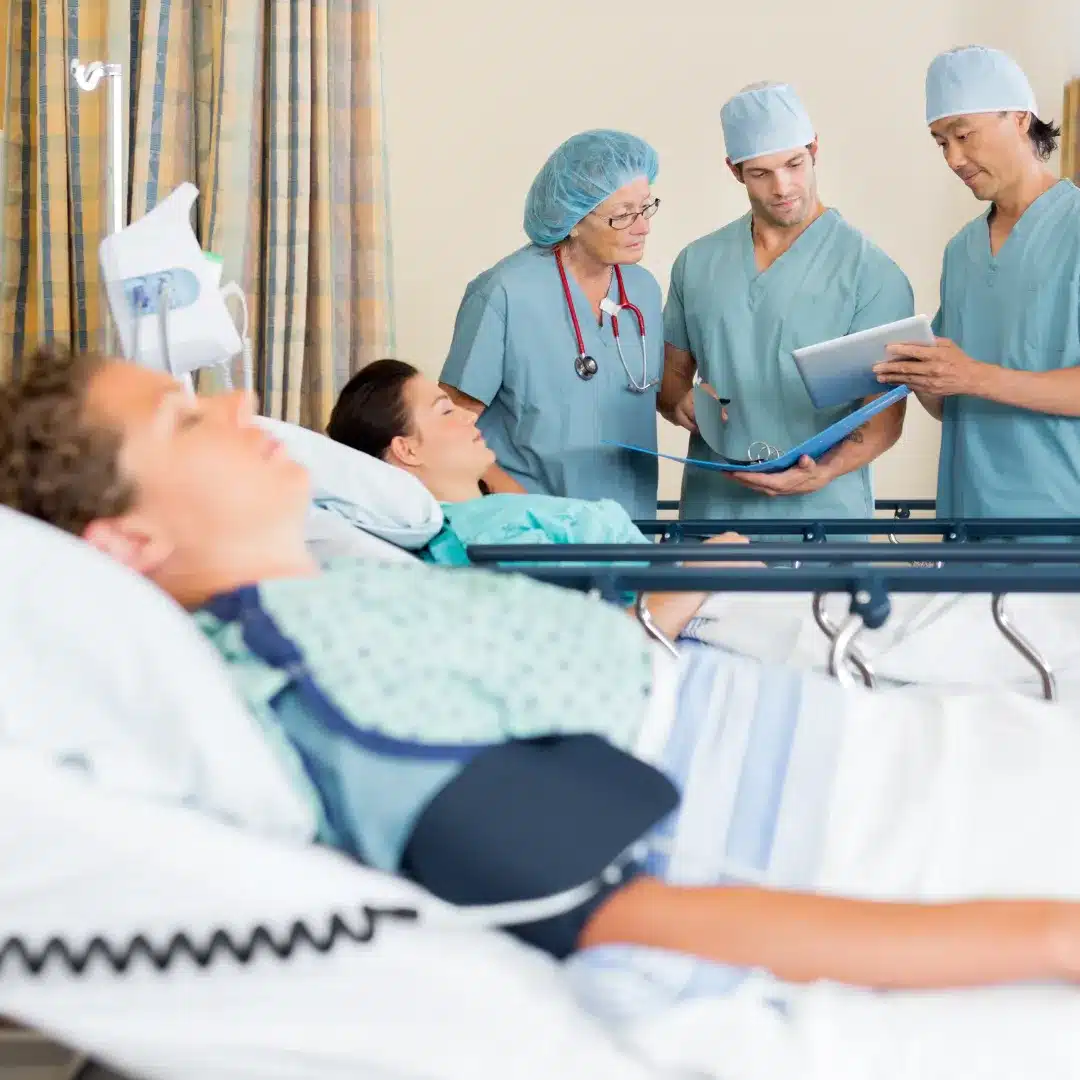 Three surgical staff in teal scrubs reviewing a tablet at a hospital bedside with patients visible.