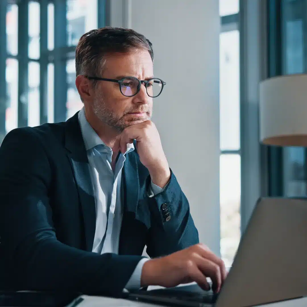Man in a dark blazer and glasses concentrating on a laptop at a desk near large office windows.