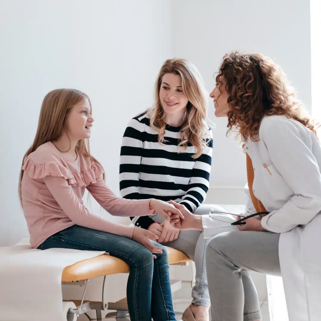 Female physician in a white coat connecting with a young patient and her mother in a pediatric exam room.