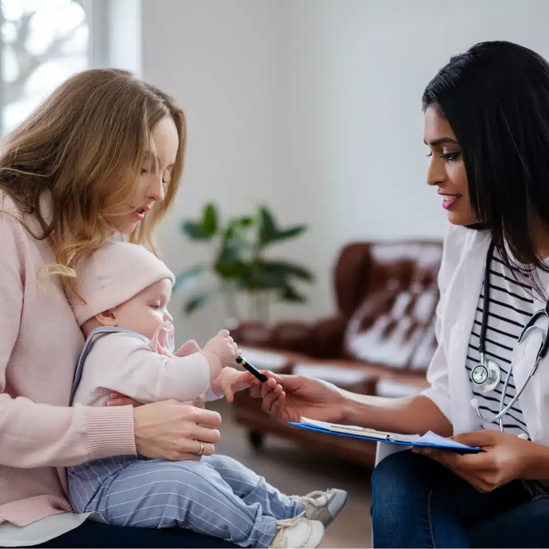 Female provider with a clipboard examining a baby held by the mother during a pediatric home visit.