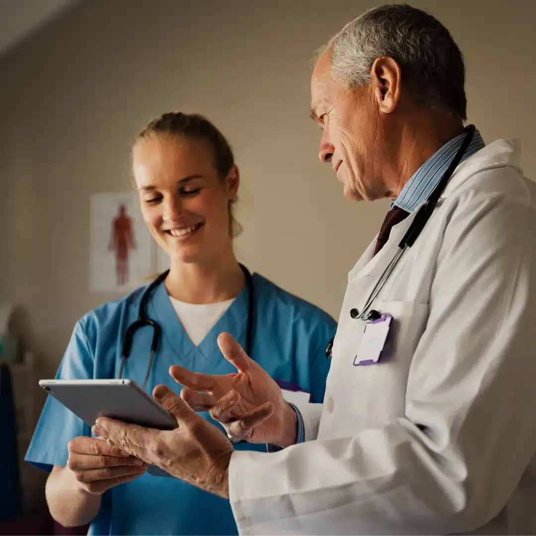 Senior physician in a white coat gesturing at a tablet held by a nurse in scrubs in an exam room.
