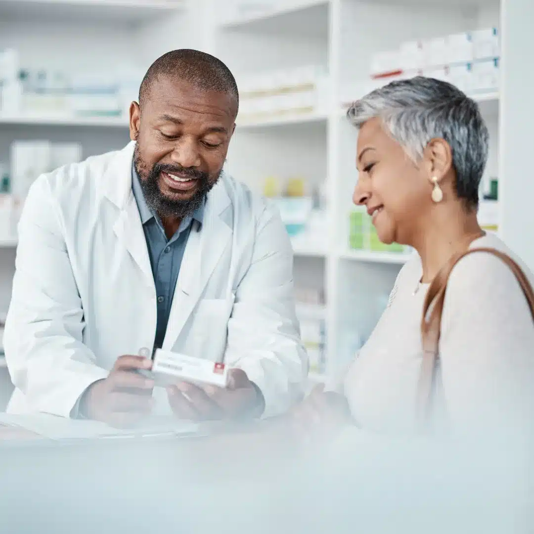 Male pharmacist in a white coat reviewing a medication box with a senior female patient at the counter.