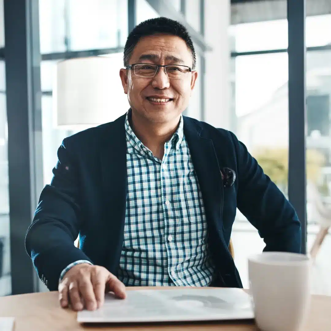 Smiling man in a dark blazer and glasses seated at a bright table with an open document and coffee.