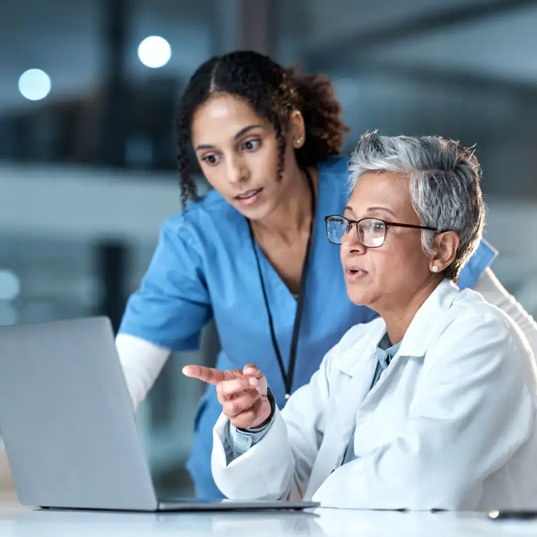 Senior physician in a white coat and nurse in scrubs reviewing content on a laptop together.