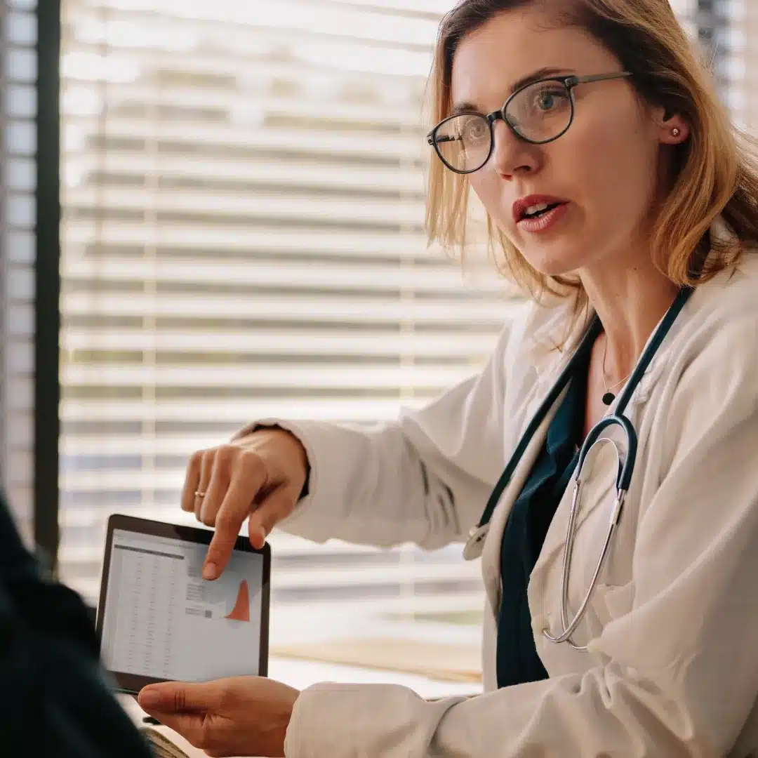 Female physician in a white coat pointing at a tablet while explaining results to a seated patient.