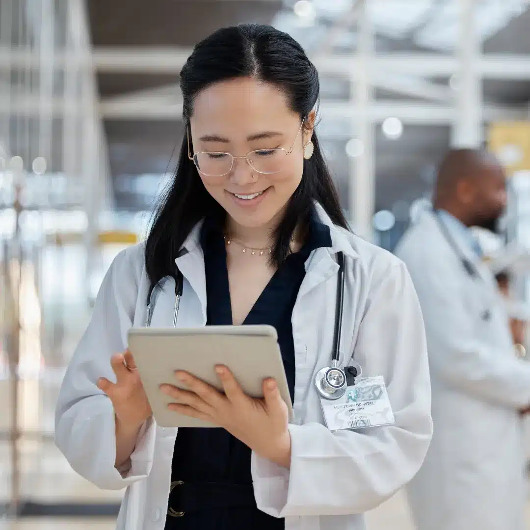 Female physician with a hospital badge smiling while reviewing a tablet in a bright clinical hallway.