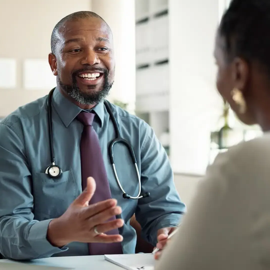 Male physician with a stethoscope smiling and gesturing while speaking with a patient at his desk.