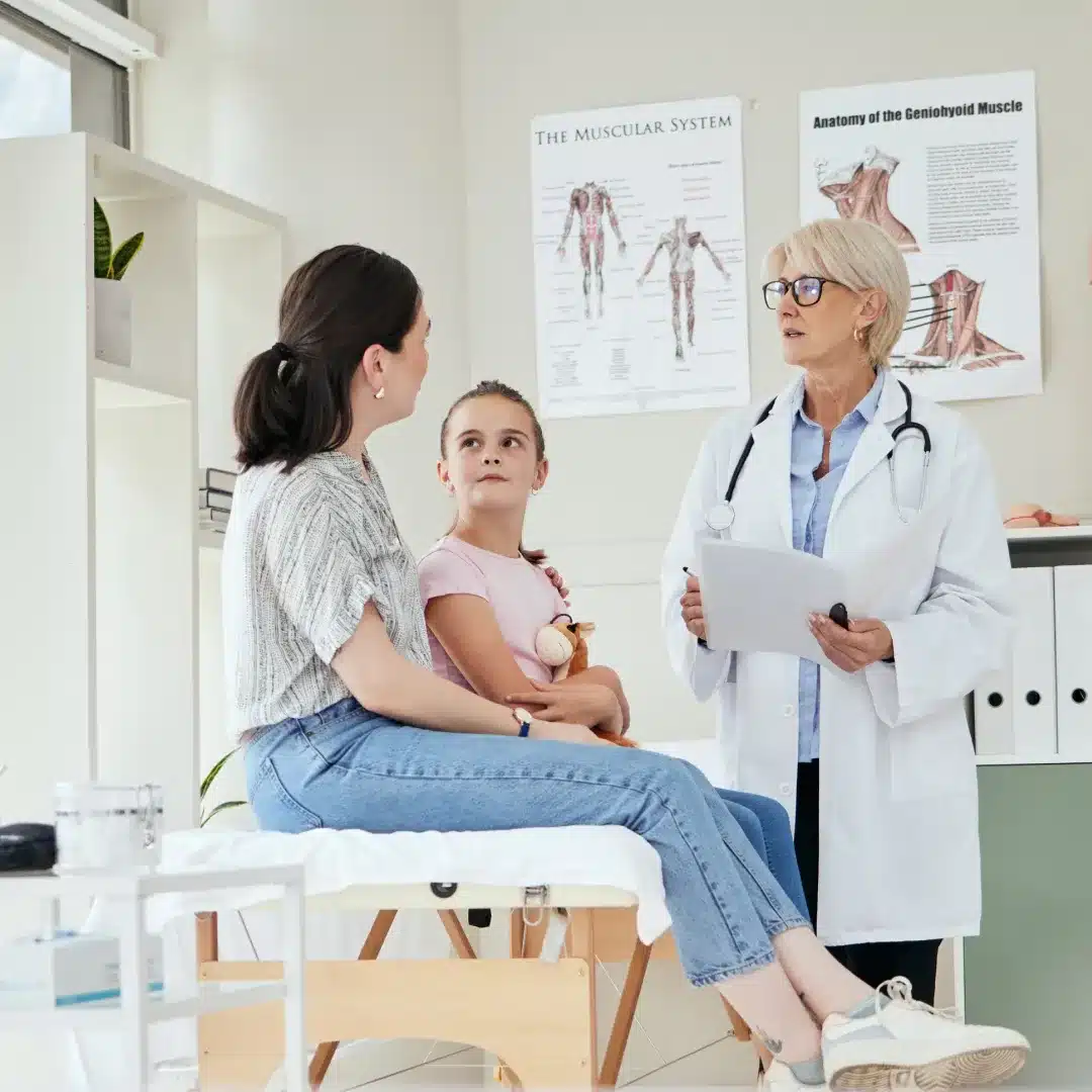 Pediatric physician in a white coat consulting with a young patient and her mother in an exam room.