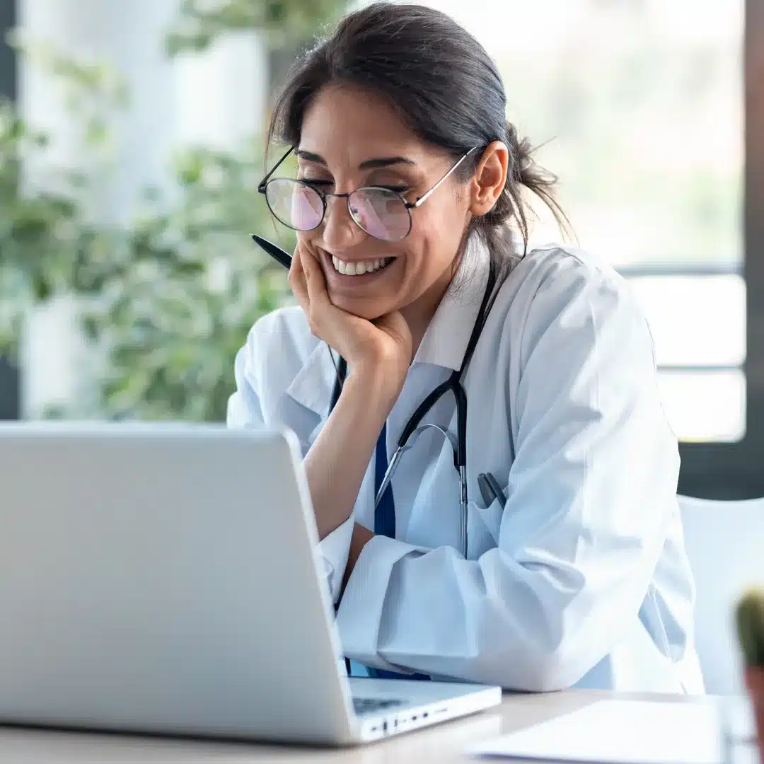 Female physician in a white coat and stethoscope smiling while reviewing records on a laptop.