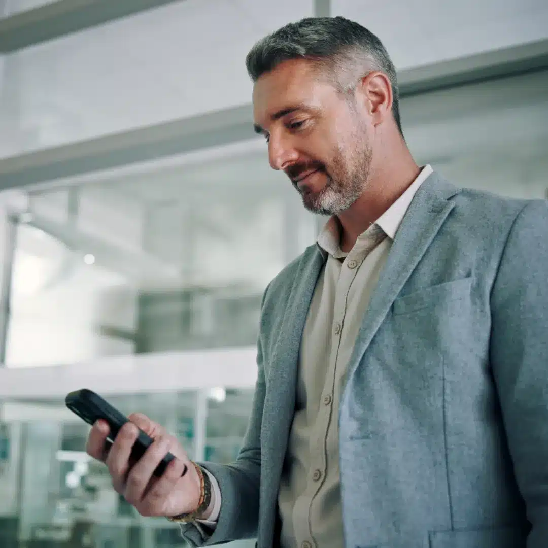 Man in a gray blazer checking his smartphone in a bright modern office hallway.