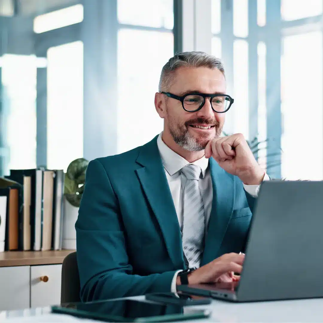 Man in a dark blazer and glasses concentrating on a laptop at a desk near large office windows.