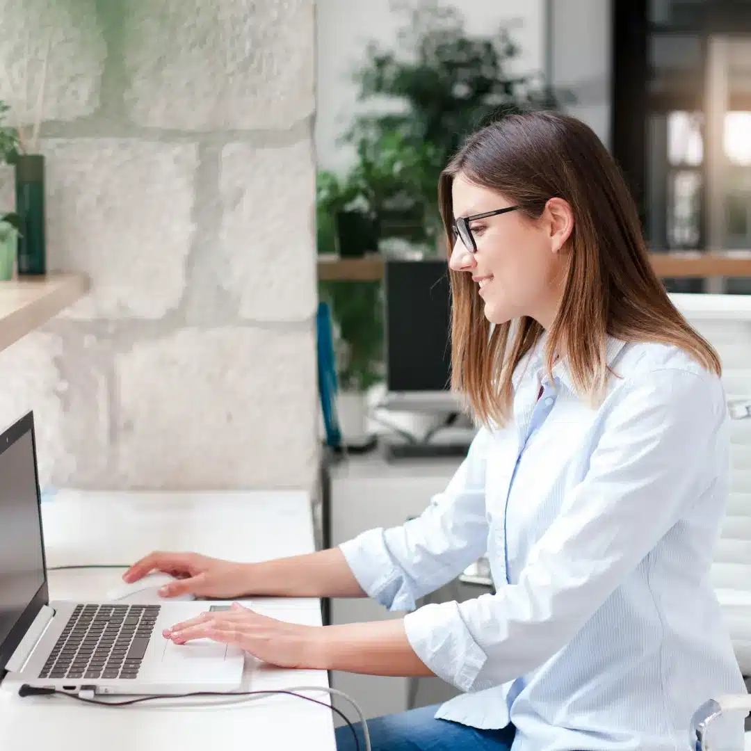 Young woman with glasses working on a laptop at a bright desk in a plant-filled office space.
