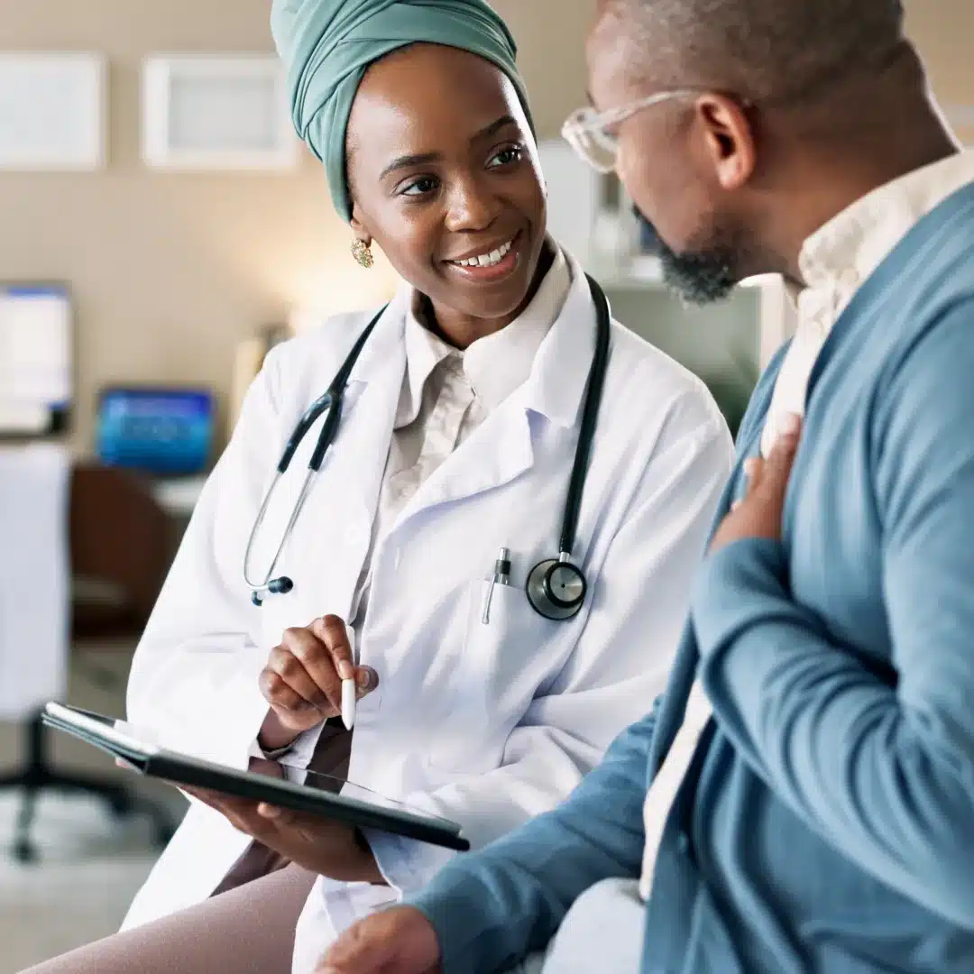 Female physician in a white coat reviewing a tablet with a patient during a clinic exam room visit.