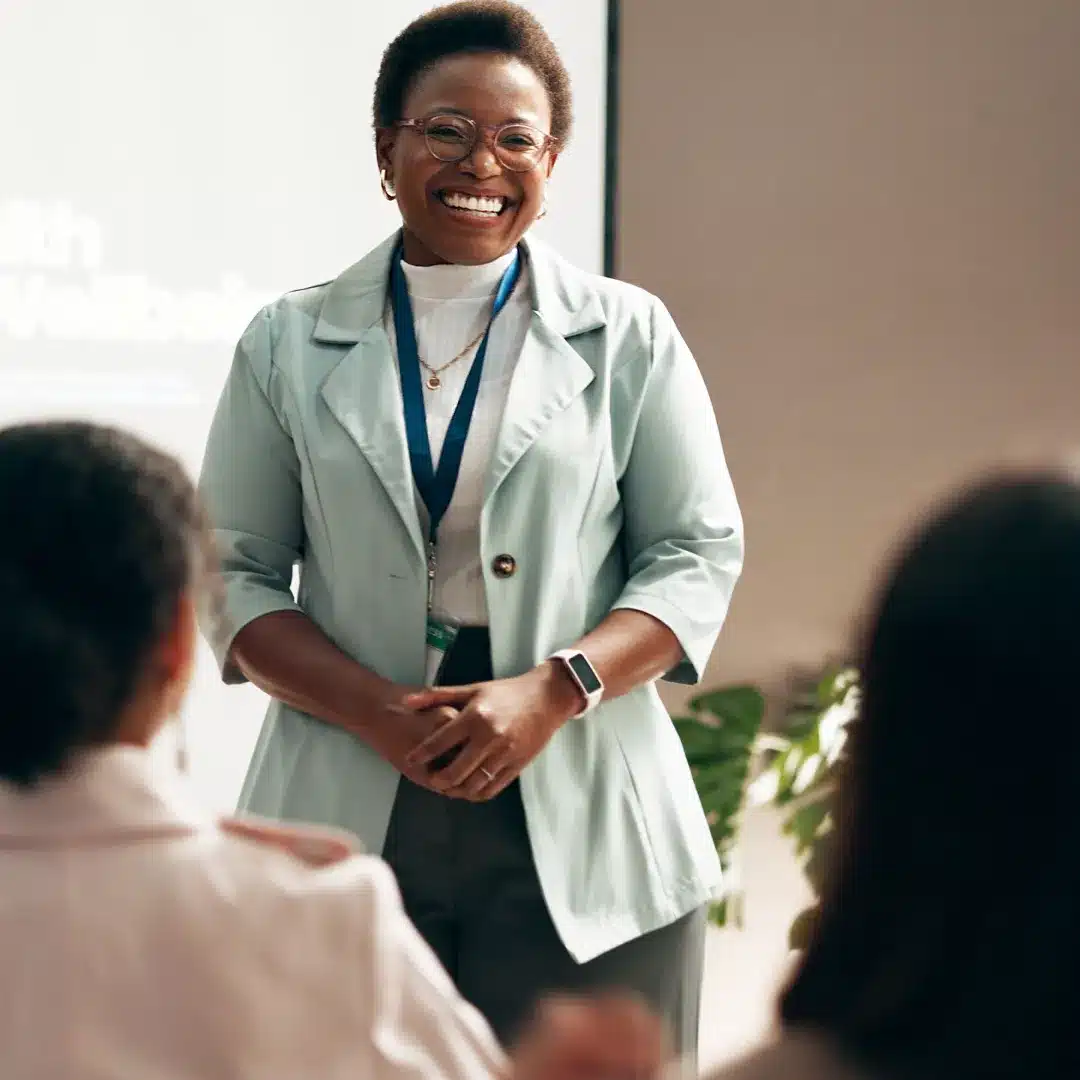Woman in a teal blazer with a lanyard presenting to a small group in a bright meeting room.