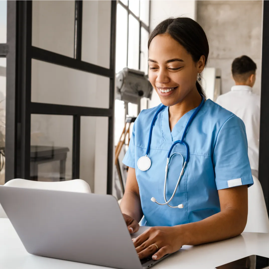 Nurse in blue scrubs and stethoscope smiling while entering data on a laptop in a medical office.