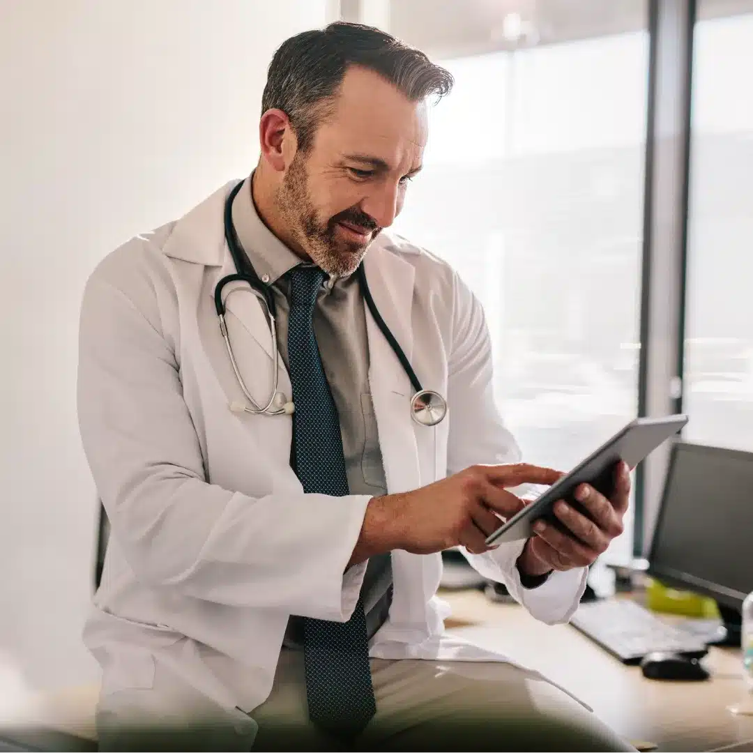 Male physician in a white coat and stethoscope reviewing a tablet in a bright medical office.
