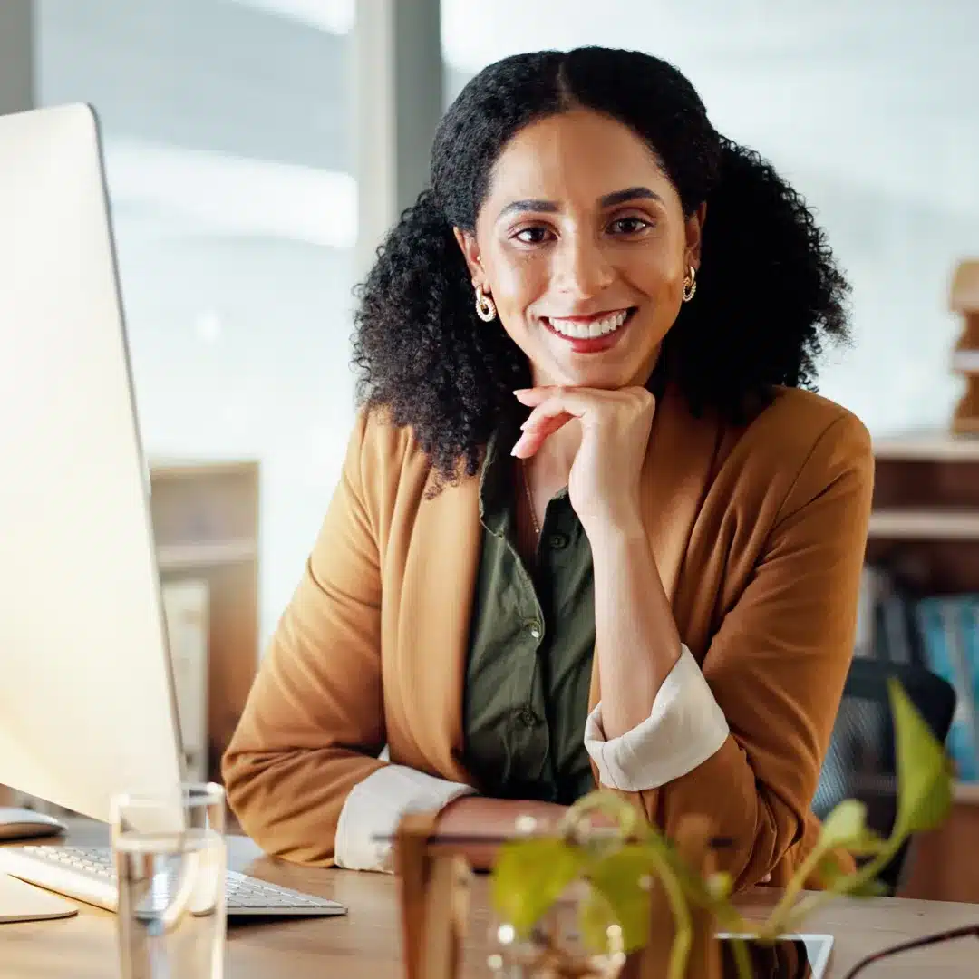 Smiling woman in a tan blazer sitting at a desktop computer in a warmly lit office setting.