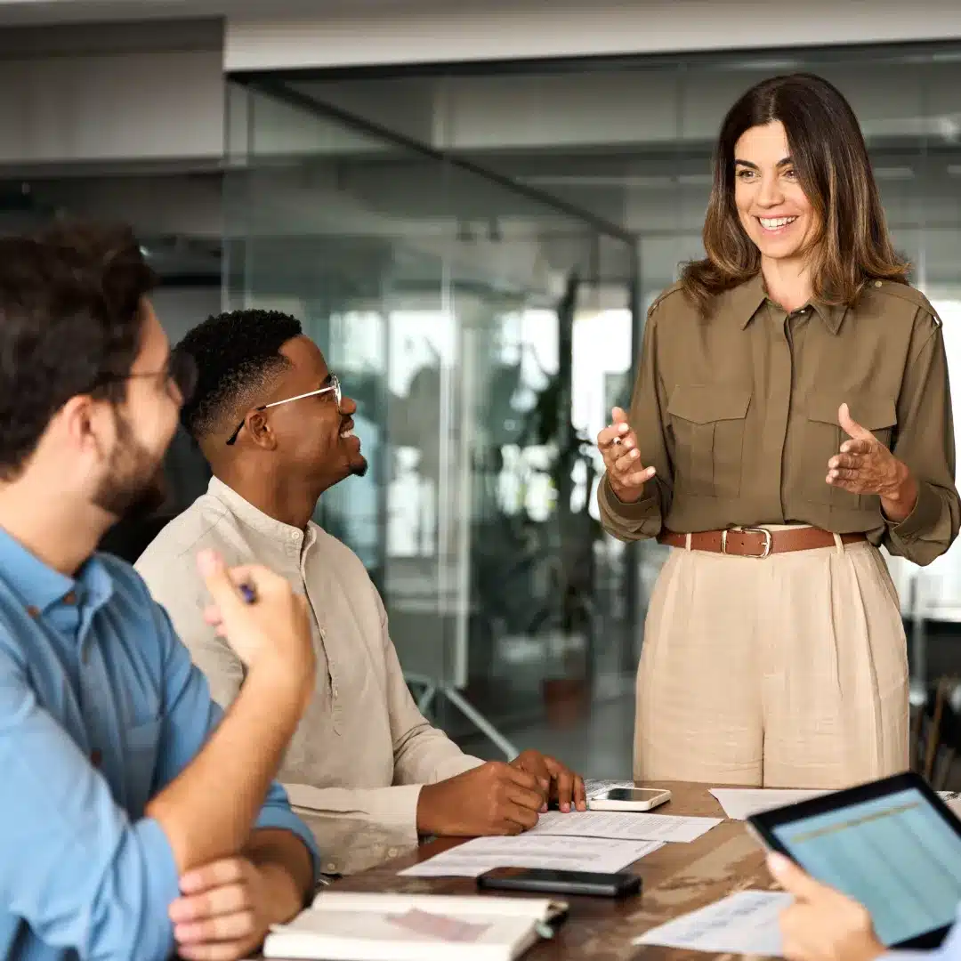 A woman stands at a conference table, engaging with three colleagues sitting around her, discussing documents and using devices.