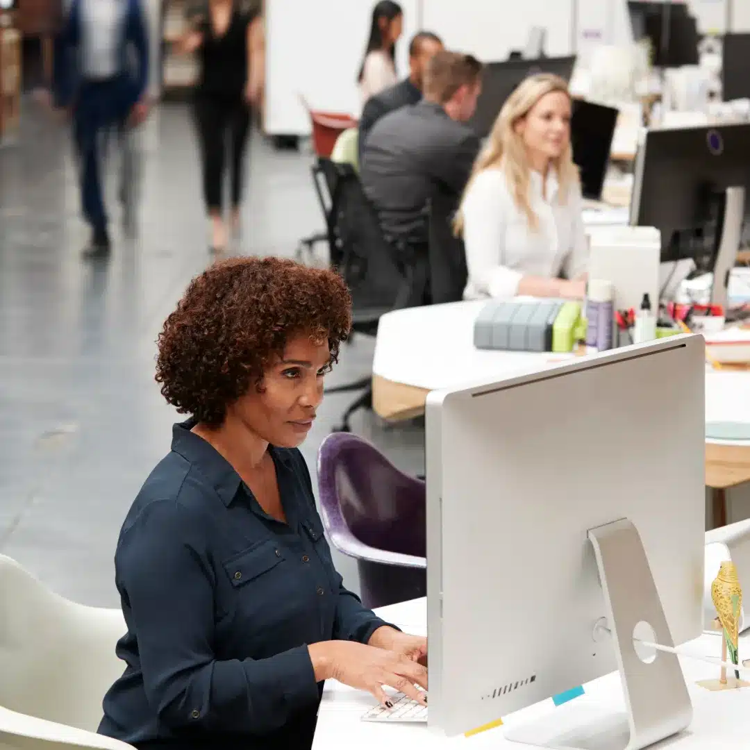 Woman with curly hair in a dark shirt working at a desktop computer in a busy open-plan office.