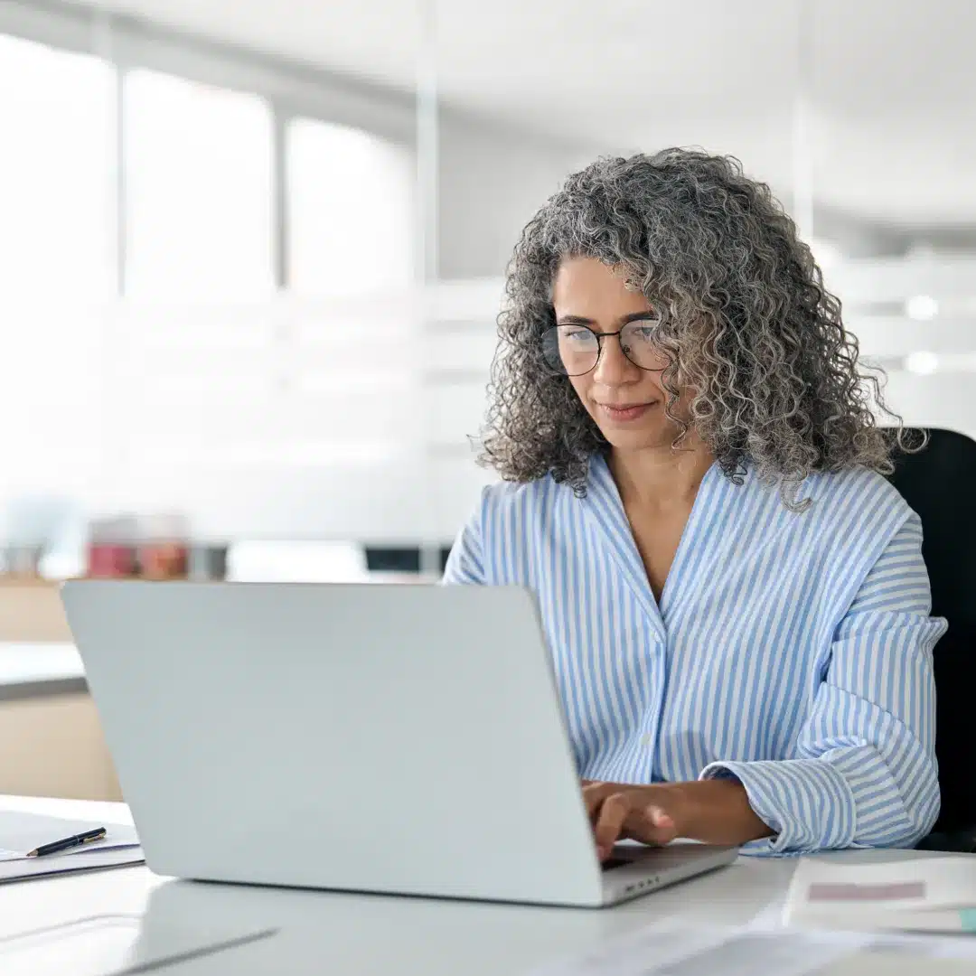 Woman with curly gray hair and glasses working on a laptop at a bright, paper-strewn office desk.