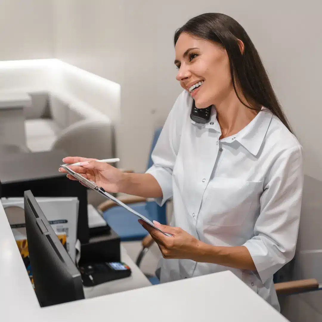 Smiling medical receptionist in a white coat holding a tablet and phone at a clinic front desk.