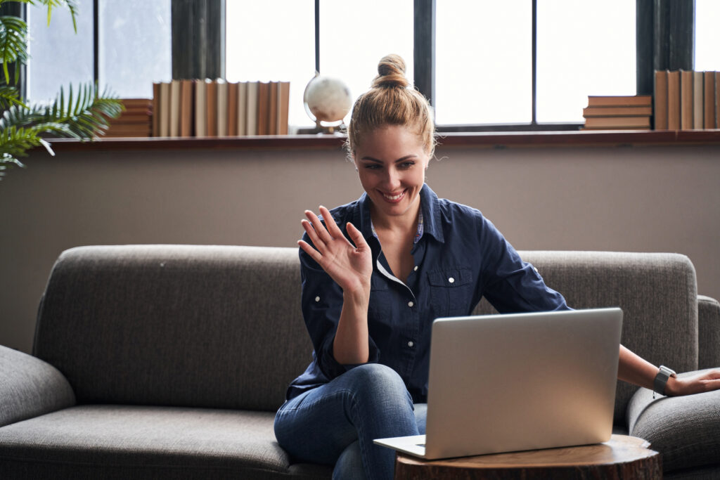 Cheerful young lady sitting on a sofa with a laptop and putting her hand up in front of a web camera