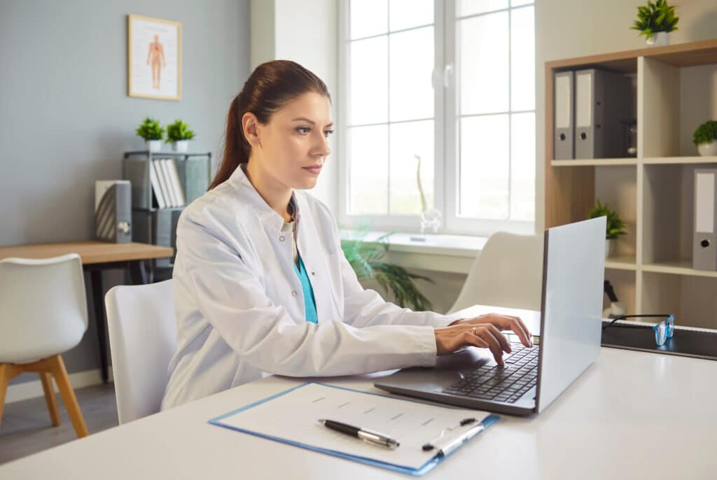 Young female doctor in white coat sits at desk and working on laptop in modern clinic office, checking patient data or medical records. Online consultations, digital health records management concept