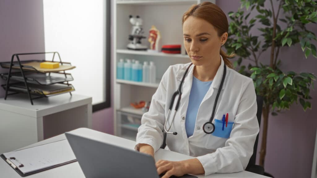 Woman working in a clinic wearing a stethoscope and white coat, focusing on a laptop amidst medical equipment and plants in a professional health care environment.