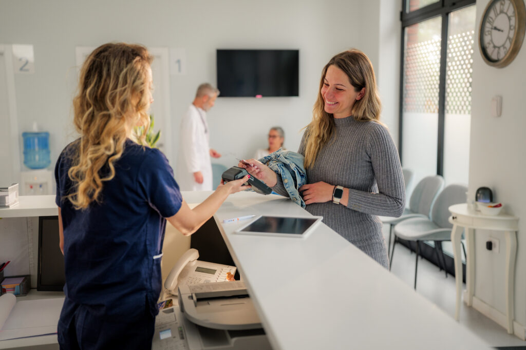 Pregnant woman is using contactless payment at the reception desk of a modern clinic, interacting with a friendly receptionist while a doctor attends to another patient in the background