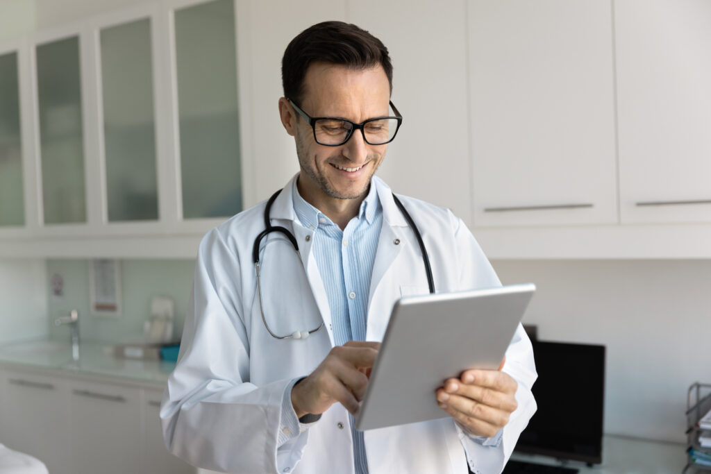 Happy male physician doctor in glasses reading medical reports on tablet, reviewing electronic lab research results, patients records, using online application on digital pad, smiling