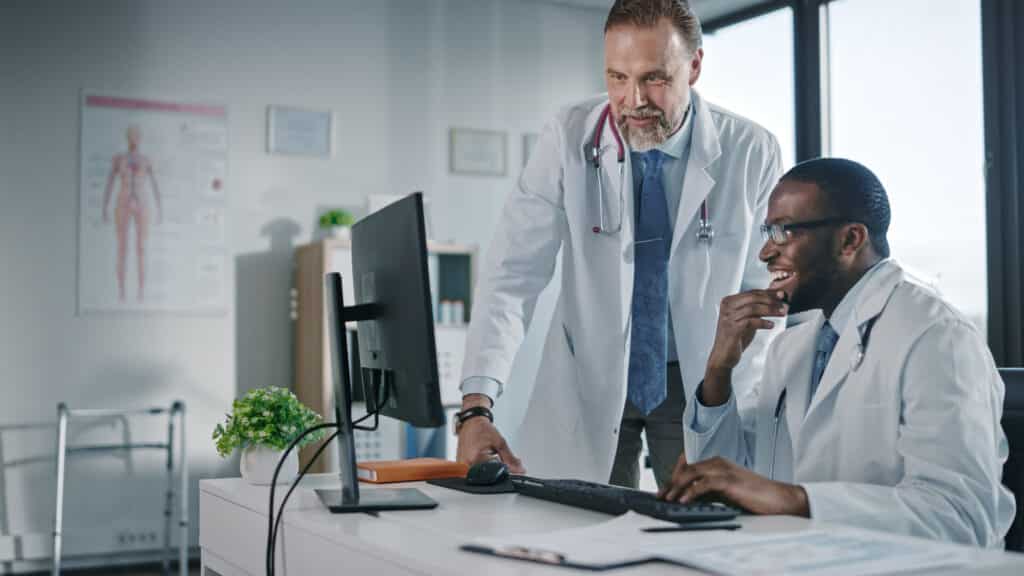 Young African American Physician is Talking to a Senior Doctor in a Medical Health Clinic. Colleagues Discuss Work on Desktop Computer in Hospital Office. Assistant Asks Medical Advice from a Doctor.