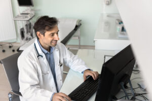 Positive Latin doctor man working at computer in hospital examination room, typing on keyboard, reading electronic records, smiling, using modern Internet technology for job in medicine