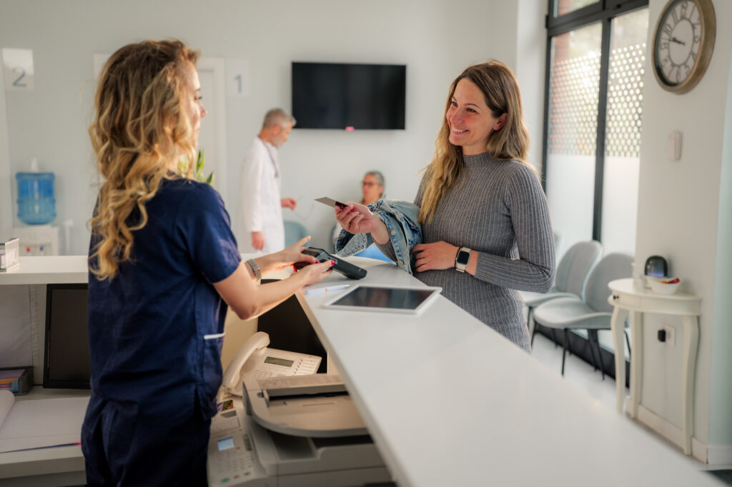 Pregnant woman is completing a contactless payment transaction at the reception desk of a modern medical clinic, interacting with the receptionist while a doctor walks in the background