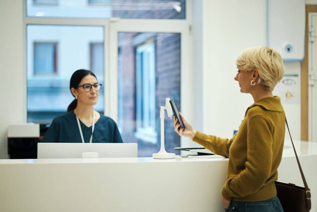 Caucasian young woman standing at reception desk in medical clinic, holding smartphone while interacting with nurse receptionist sitting behind counter in modern hospital setting
