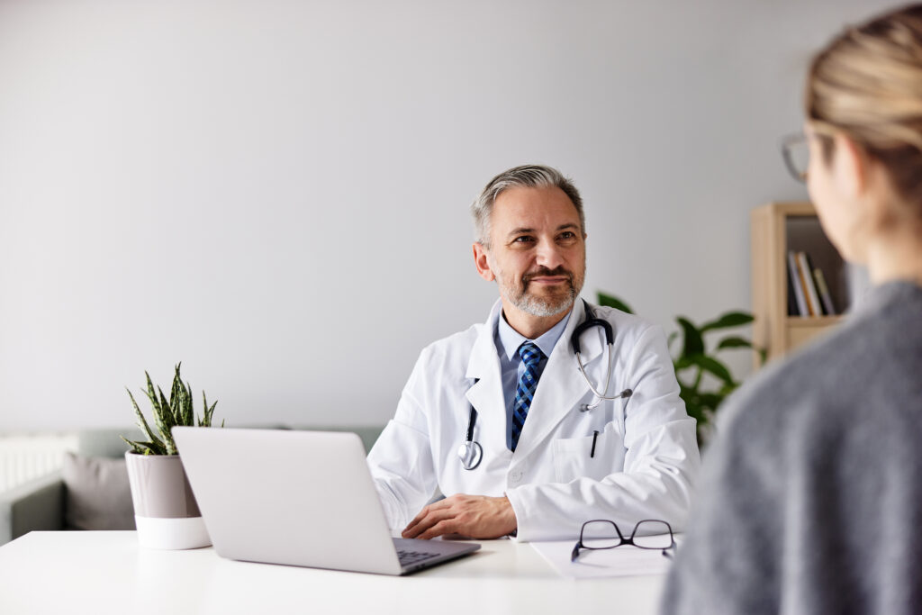A male doctor sitting in front of a laptop with glasses on a work desk, listening to a female patient, talking about health care.