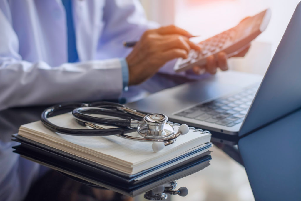 Female doctor or practitioner using calculator and work on laptop computer with medical stethoscope and notebookon the desk at clinic or hospital. Medical healthcare costs ,fees and revenue concept.